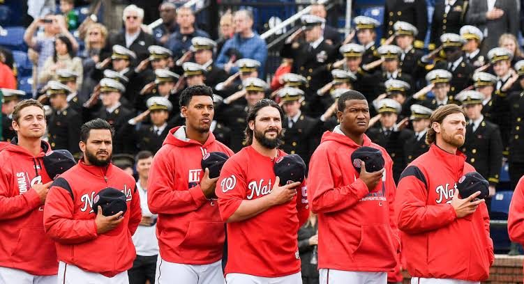 Nationals players with caps over their hearts during the anthem, rows of midshipmen saluting in the stands behind them