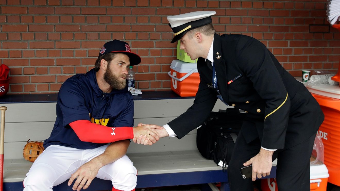 Bryce Harper sitting in the dugout in a Navy pullover, shaking hands with a midshipman in dress blues