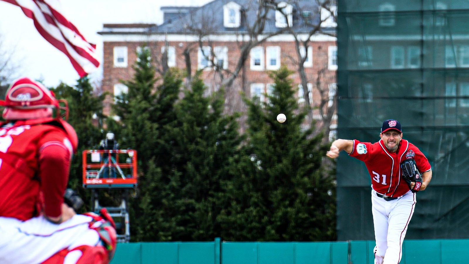 Max Scherzer delivering a pitch at Terwilliger Brothers Field with Bancroft Hall behind him